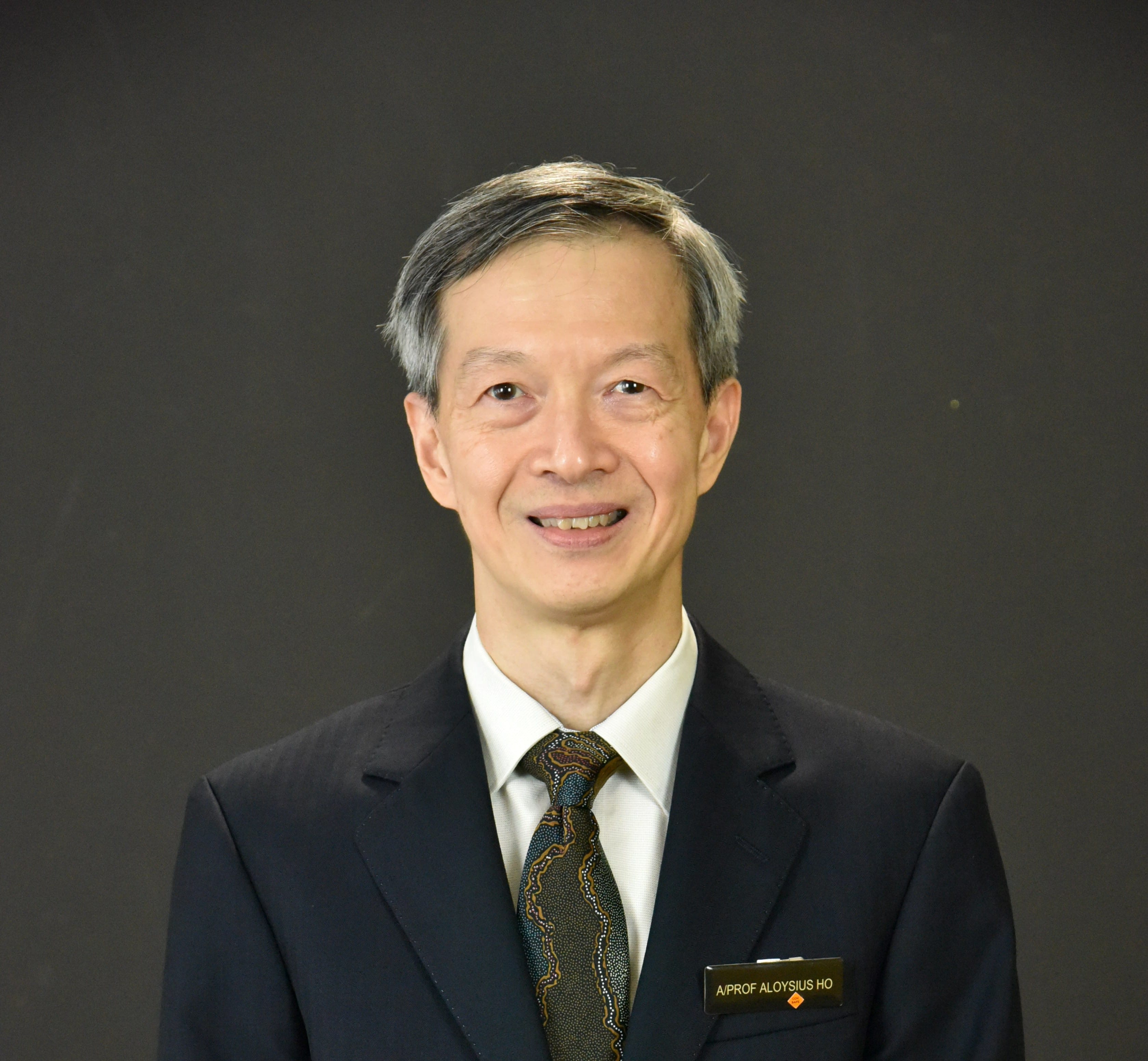 Smiling man in a dark suit and patterned tie, standing against a plain dark background.